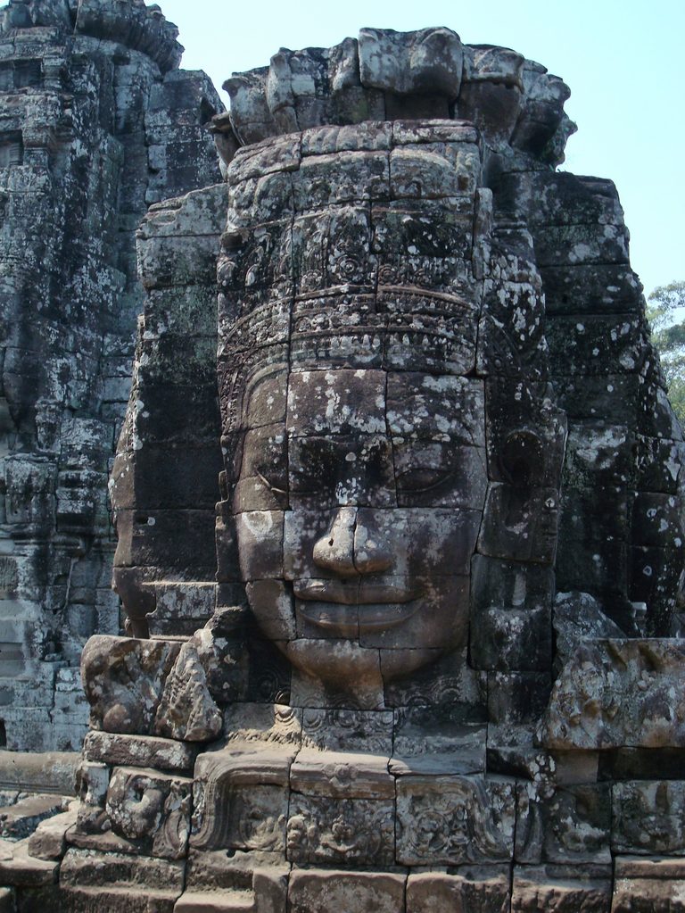 A serene smiling stone face carved on a Bayon tower against blue sky.