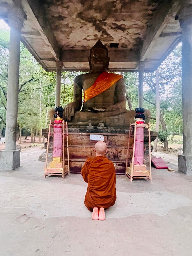 A robed monk kneeling in prayer before a forest Buddha shrine near Siem Reap