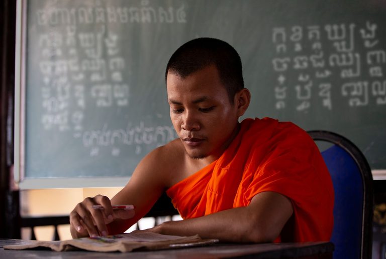 An adult monk in orange robes writes at a desk below a Khmer-chalked blackboard.