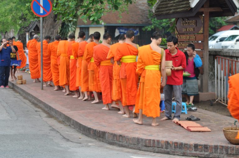 A line of barefoot monks in saffron robes collecting morning alms along a street.