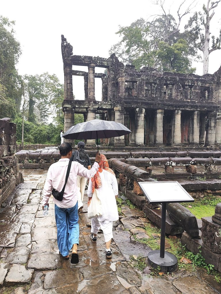 Two travellers under one umbrella walking toward Preah Khan temple in the rain